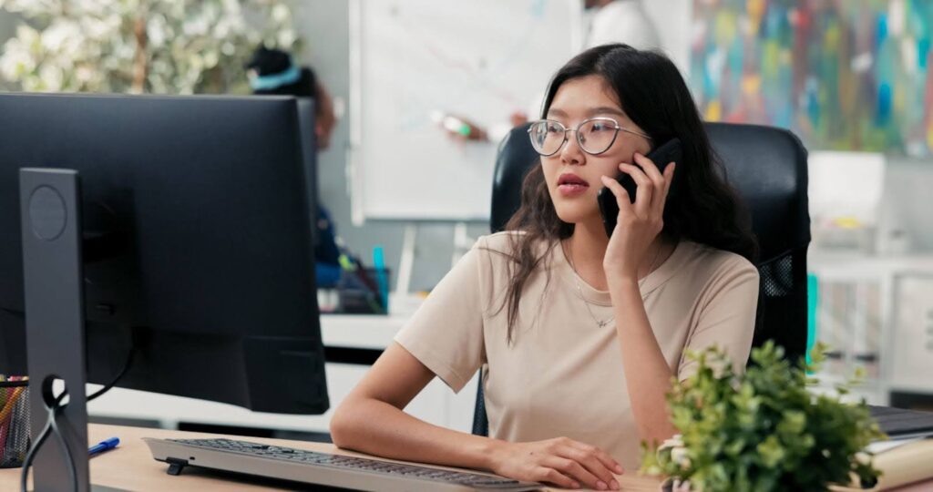 A female Asian outsourced appointment scheduler in the office, sitting in front of her computer and talking on the phone