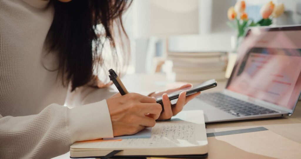 A close-up of an outsourced scheduler looking at her smartphone and taking notes in a notebook
