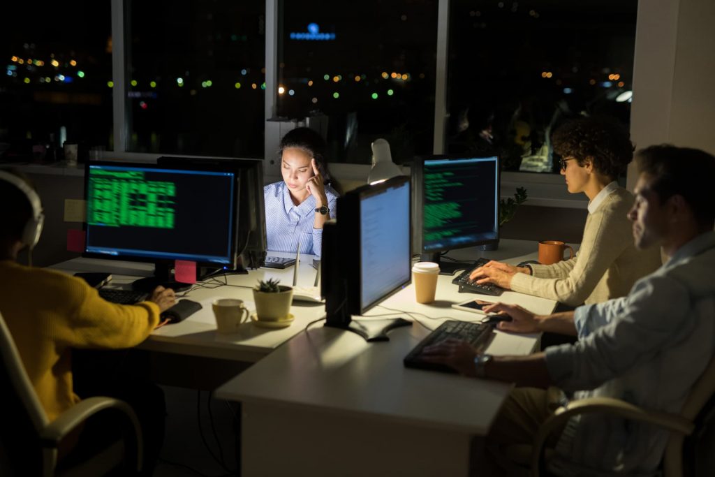 A team of programmers working late at night, focused on code displayed on their laptops